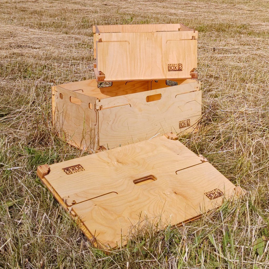 Klappboxen im Größenvergleich. Eine Kleine Holzbox steht auf der Großen, davor liegt eine Große faltbare Kiste aus Holz, die zusammengefaltet ist.
